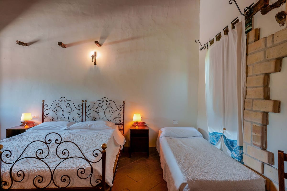 Une chambre avec deux lits, chacun doté d’une tête de lit et d’une table de chevet avec une lampe. La chambre a un mur en briques et une fenêtre avec des rideaux.