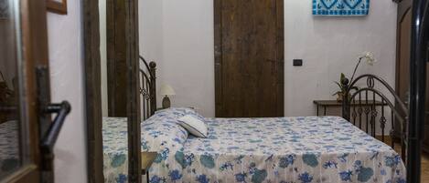A bedroom with two beds, a wooden door, a blue patterned wall hanging, and a small table with a vase.