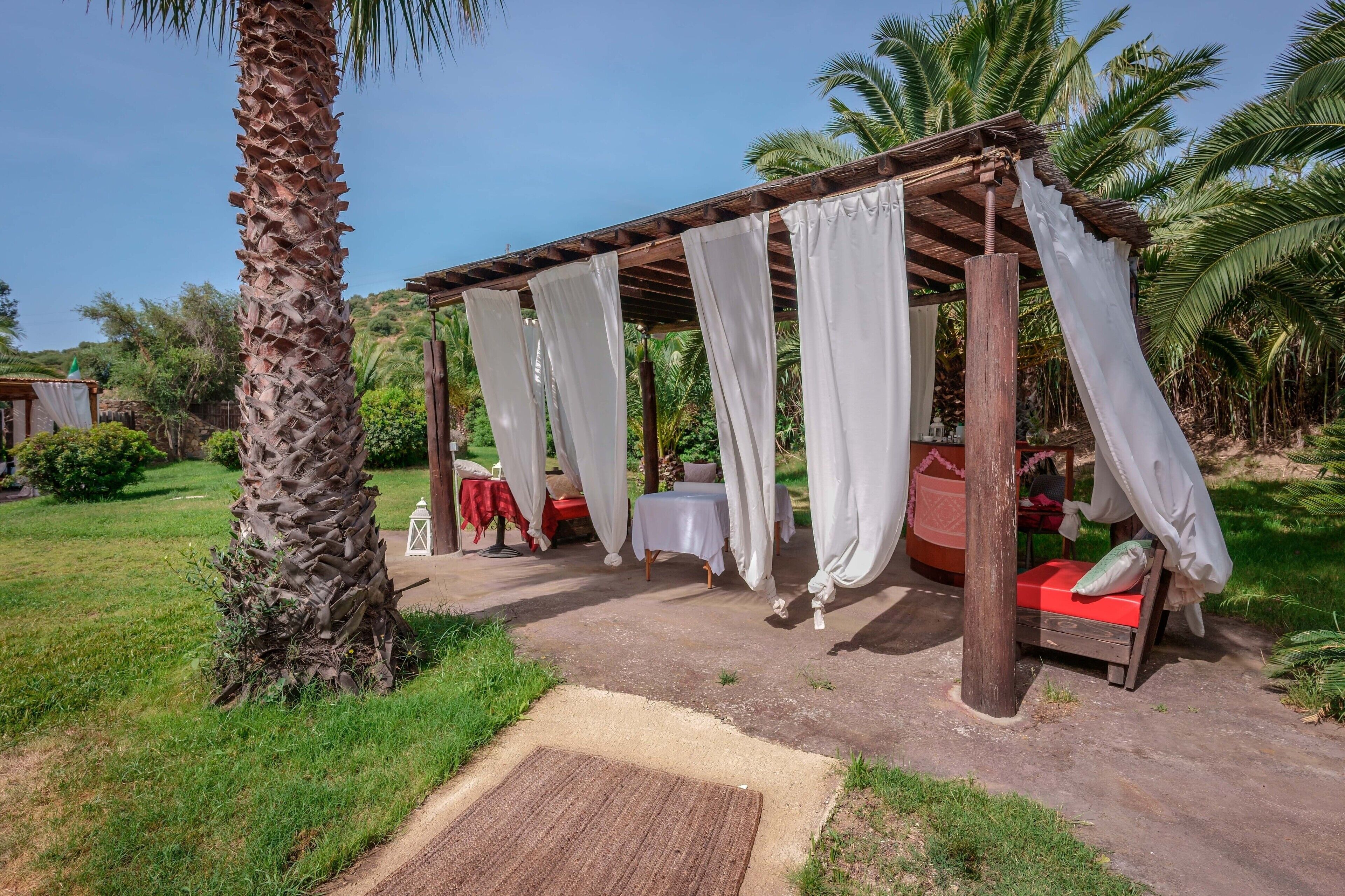 Un espacio de estar al aire libre rústico con una pérgola de madera, cortinas blancas y un banco rojo.