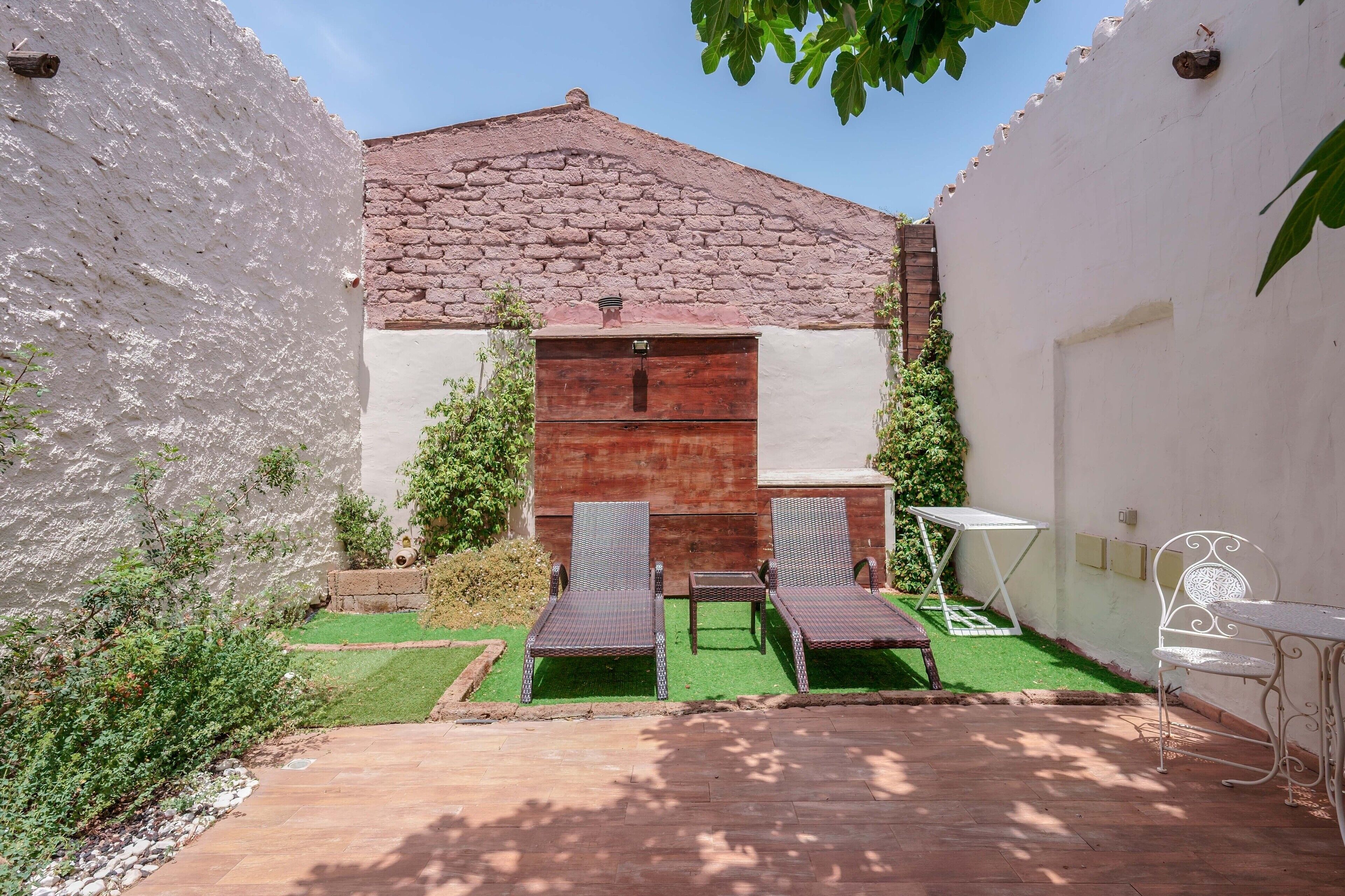 Une cour avec un mur en briques, une porte en bois, deux chaises en osier, une petite table et une chaise blanche.