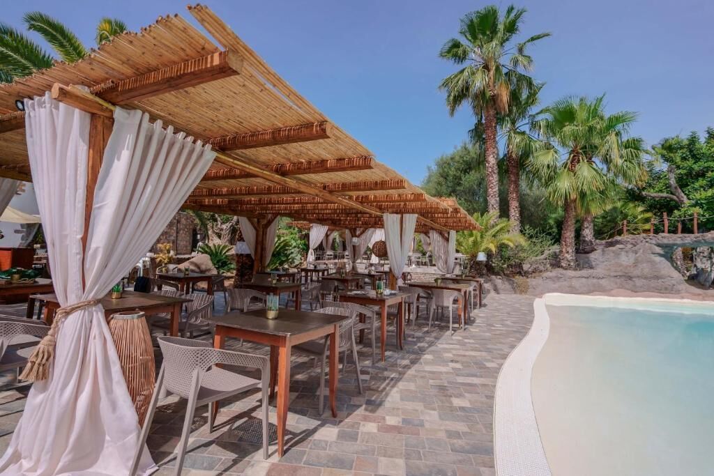Un área de comedor al aire libre con mesas y sillas de madera, cubierta por sombrillas de paja, y una piscina cerca de la zona de estar.