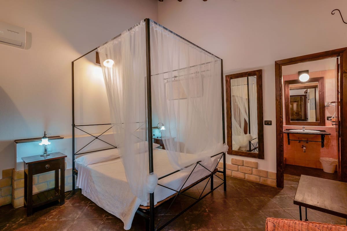 Un lit à baldaquin, une table de chevet avec une lampe, un miroir et une salle de bain avec un lavabo et un miroir.