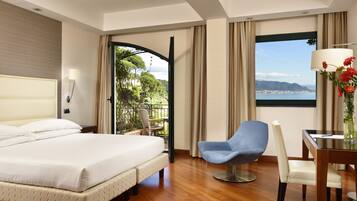 A hotel room with a bed, a desk with a chair, a view of the city and mountains, and a balcony with plants.