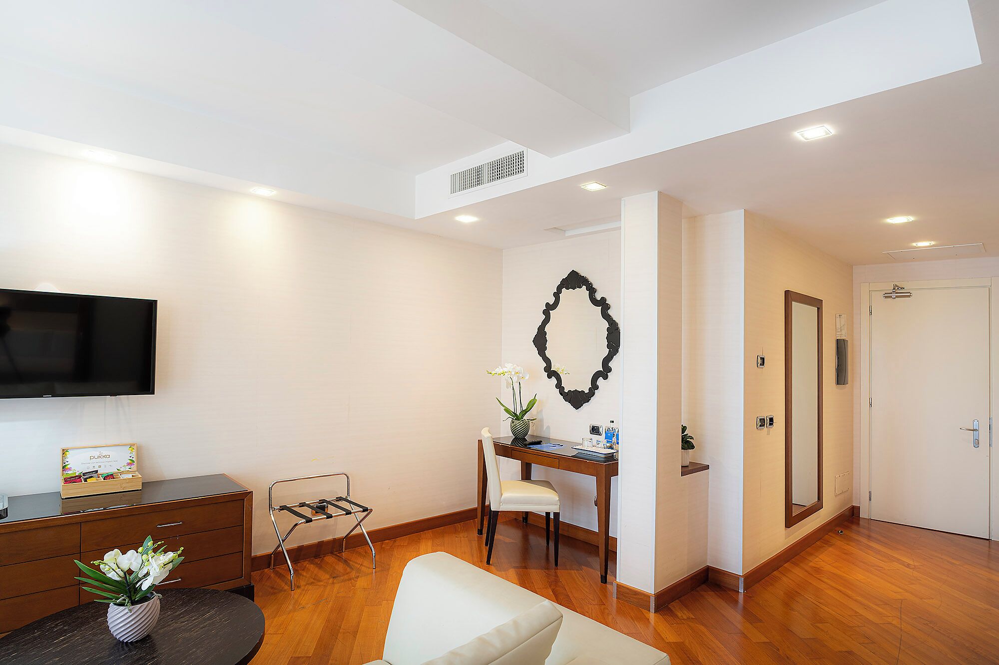 Una habitación de hotel moderna con una televisión de pantalla plana, un escritorio con una silla, una mesita con un jarrón de flores y un espejo grande en la pared.