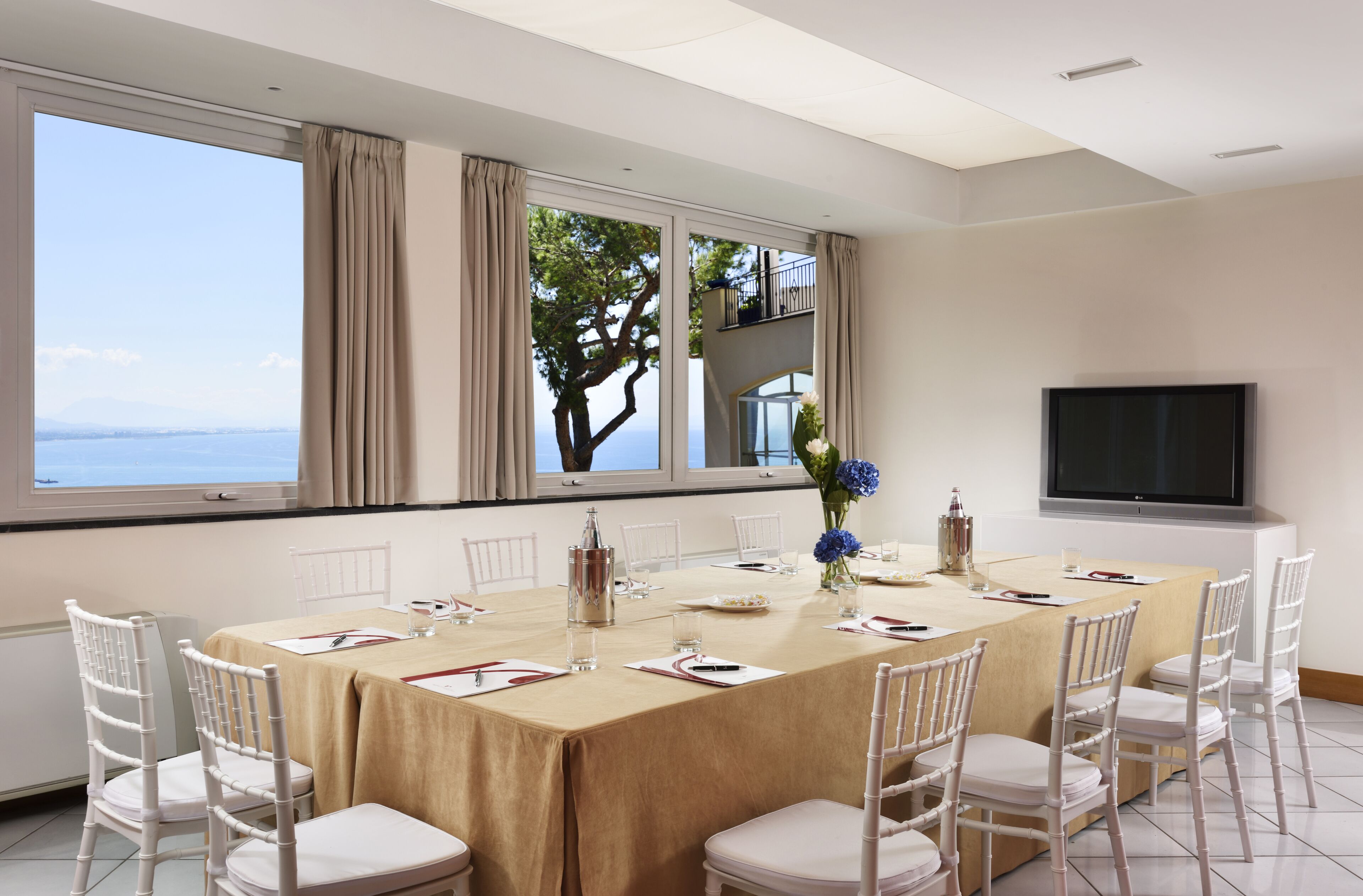 A conference room with a large table, white chairs, a TV, and a view of the sea.