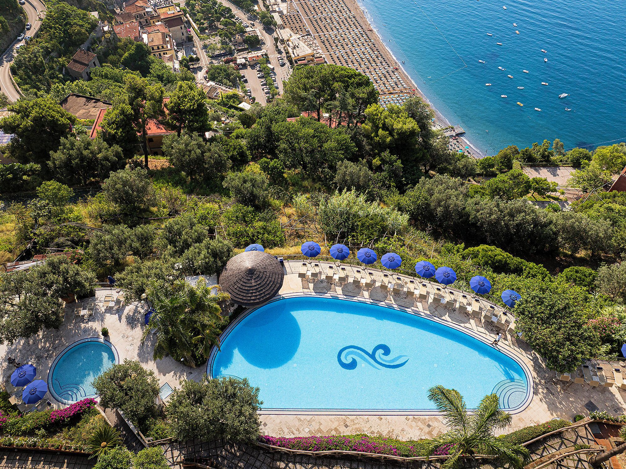 Un resort con una amplia piscina rodeada de sillones de playa y sombrillas, una playa con coches aparcados y vistas al mar.