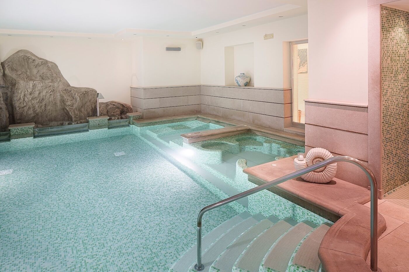 A spa pool with steps leading into it, a large rock wall, and a tiled wall with a vase.