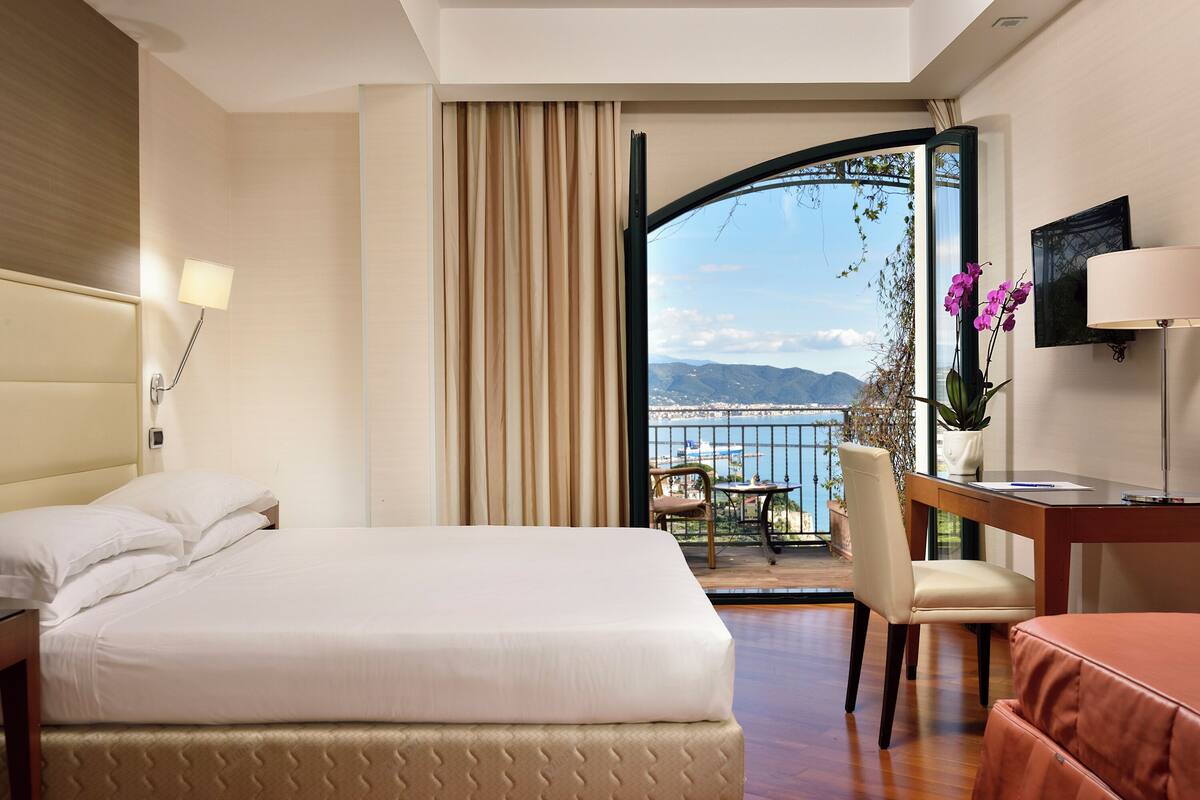 A hotel room with a bed, a desk, a chair, and a view of the sea.