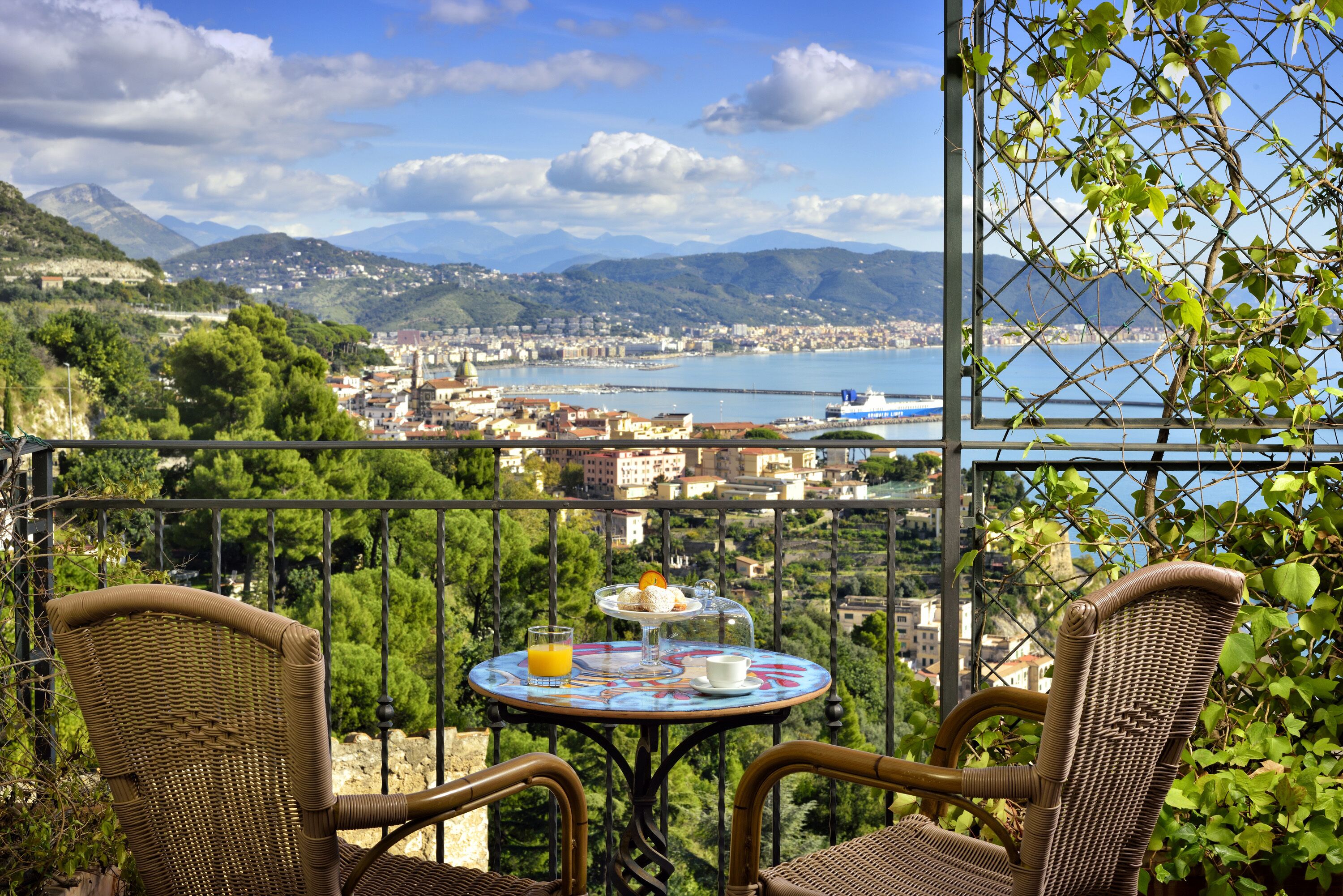 A balcony with wicker chairs, a table with a glass of orange juice, and a view of a coastal town with mountains in the background.