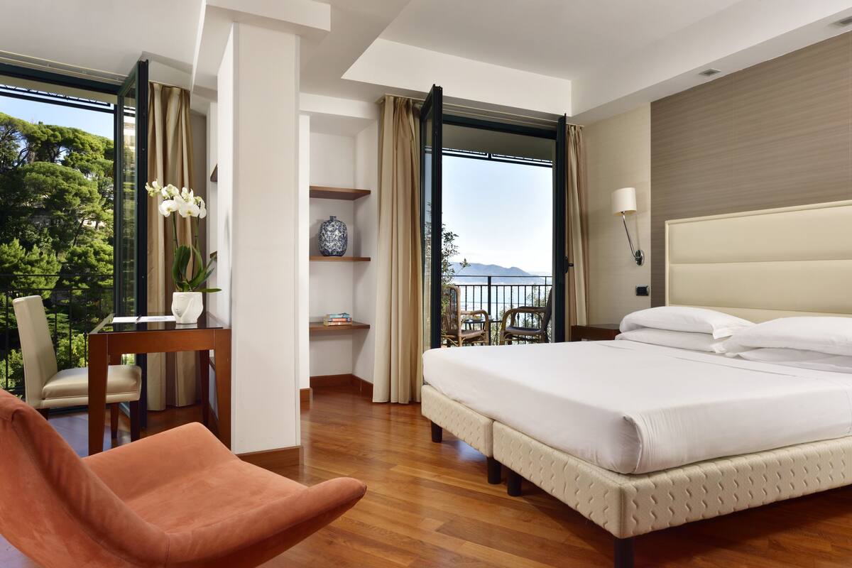 A hotel room with a large bed, a desk with a chair, a small table with a vase, and a view of the outdoors.