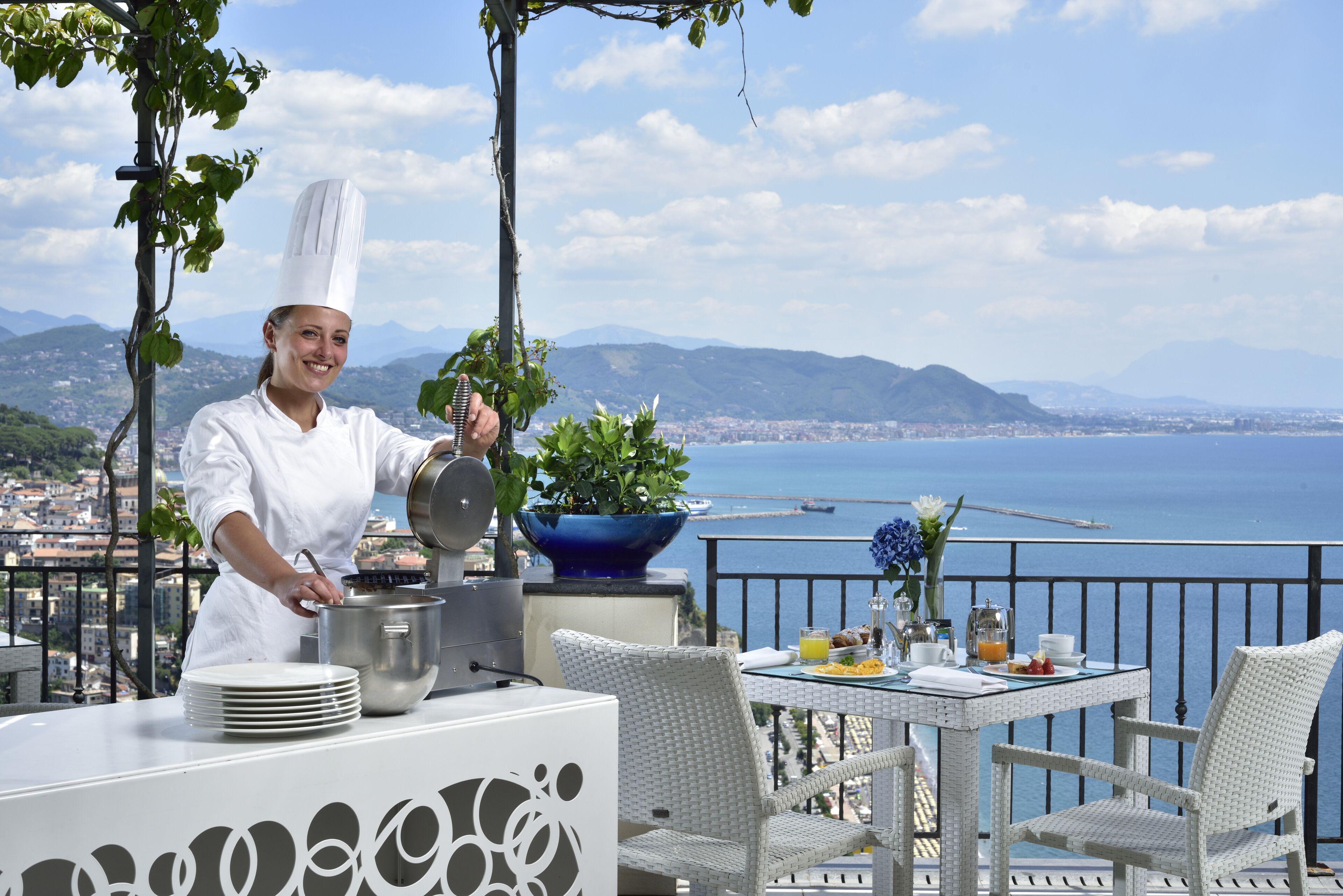 Un chef prepara comida en un balcón con vistas panorámicas al mar y las montañas.