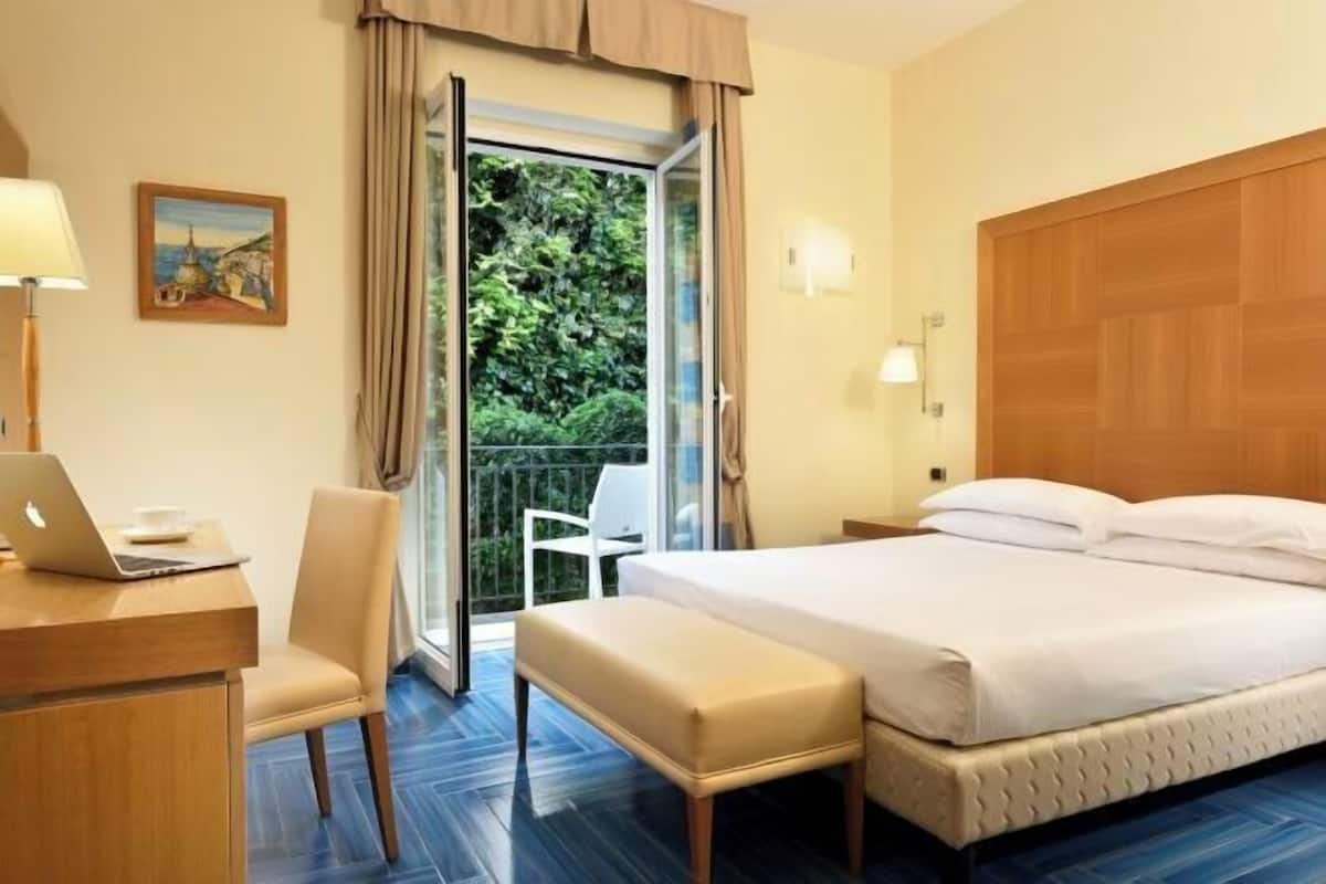 A hotel room with a bed, a desk with a laptop, a chair, a bench, a window with curtains, and a view of greenery outside.