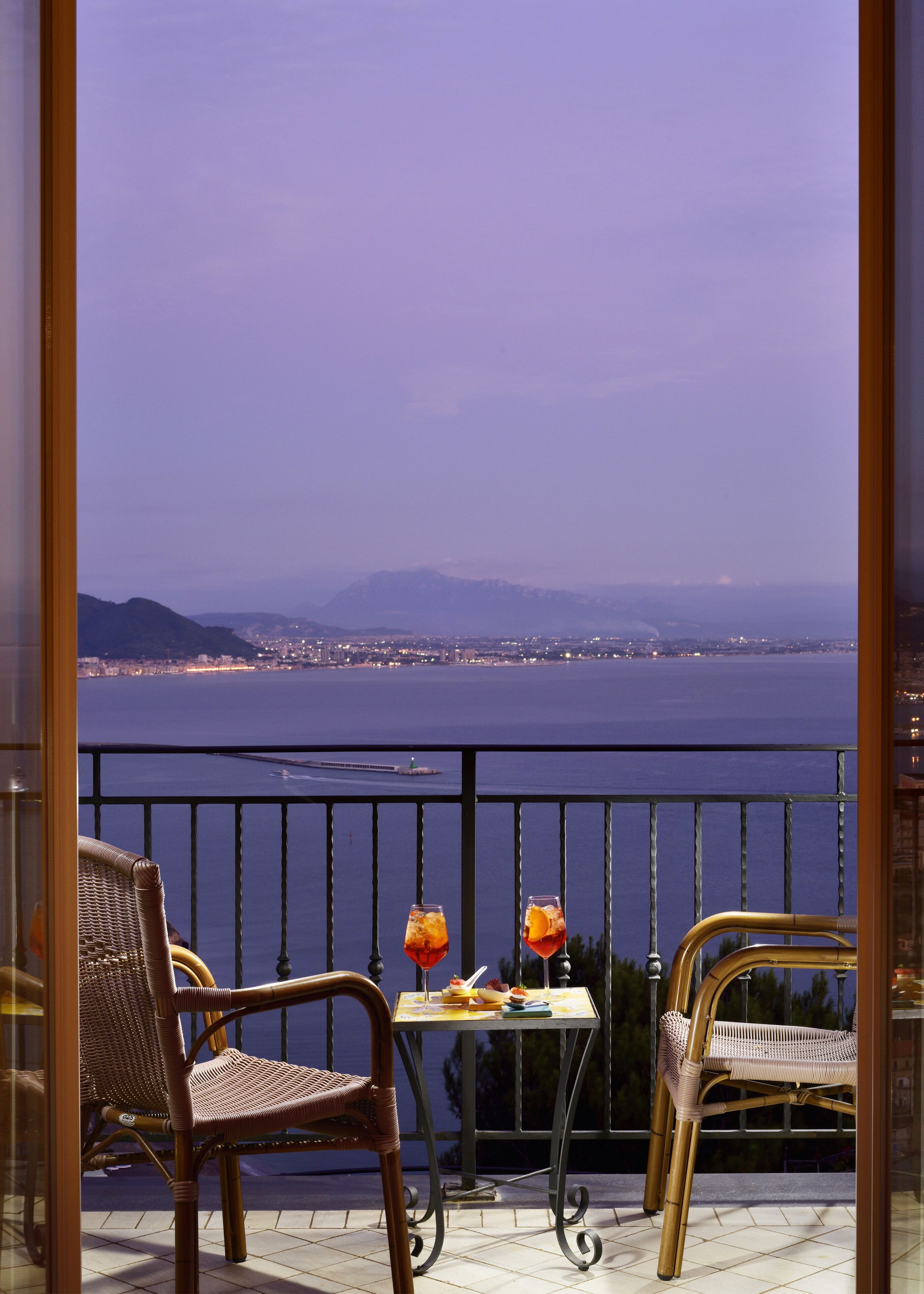 Een balkon met rieten stoelen en een tafel, met uitzicht op een waterpartij en een verafgelegen stadsbeeld.