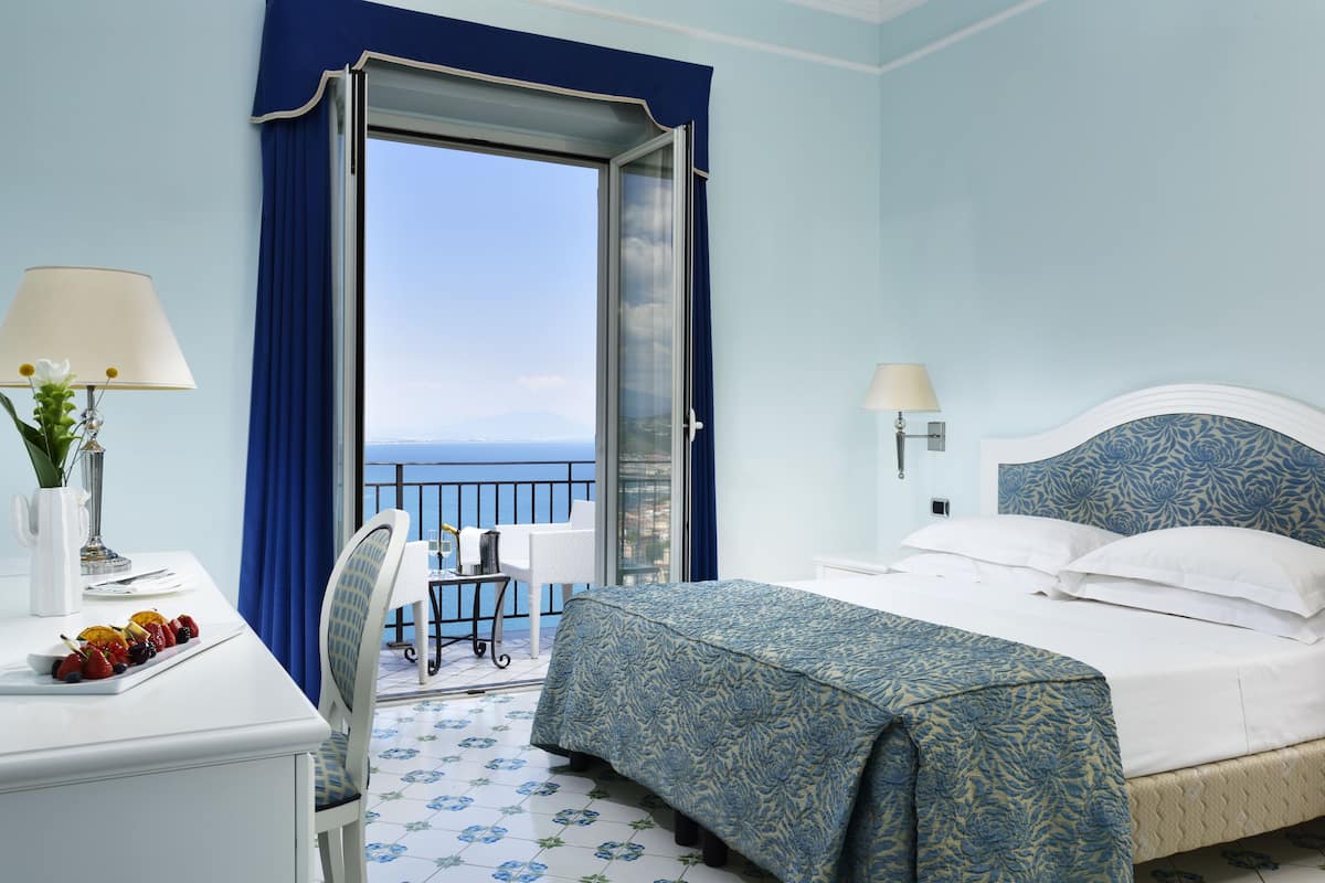 A hotel room with a bed, a desk with a lamp, a chair, and a view of the sea.