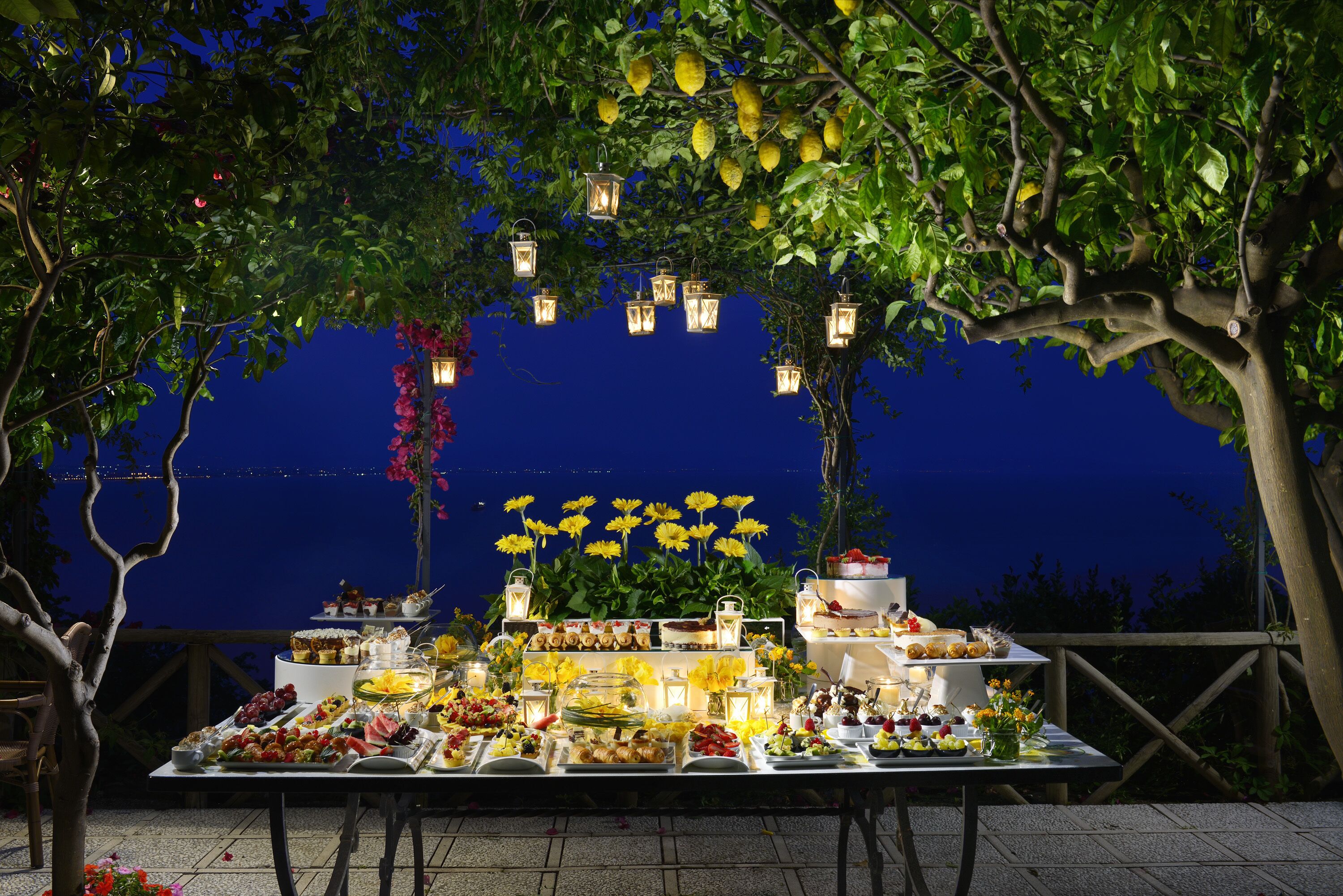 An outdoor dining area with a table set for a meal, surrounded by trees and lanterns.