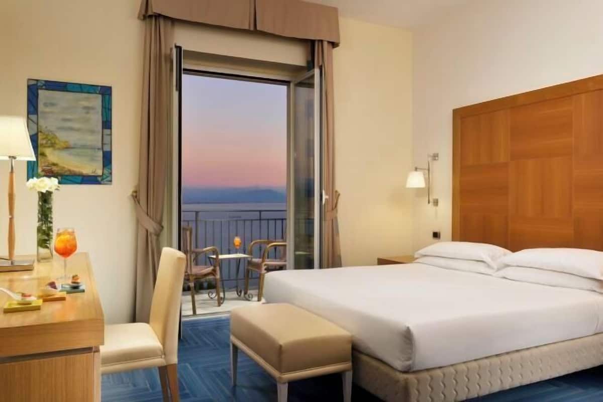A hotel room with a bed, a desk with a lamp, a chair, a small table with a drink, and a view through a glass door.