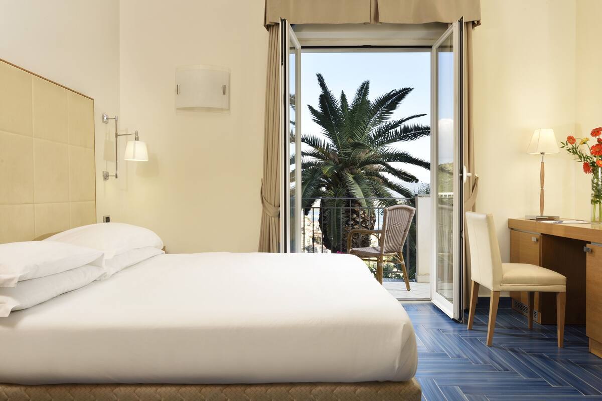 A hotel room with a bed, a desk, a chair, a lamp, and a view of palm trees through a glass door.