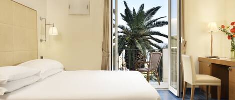 Habitación de hotel con cama, escritorio, silla, lámpara y vista a palmeras a través de una puerta de vidrio.