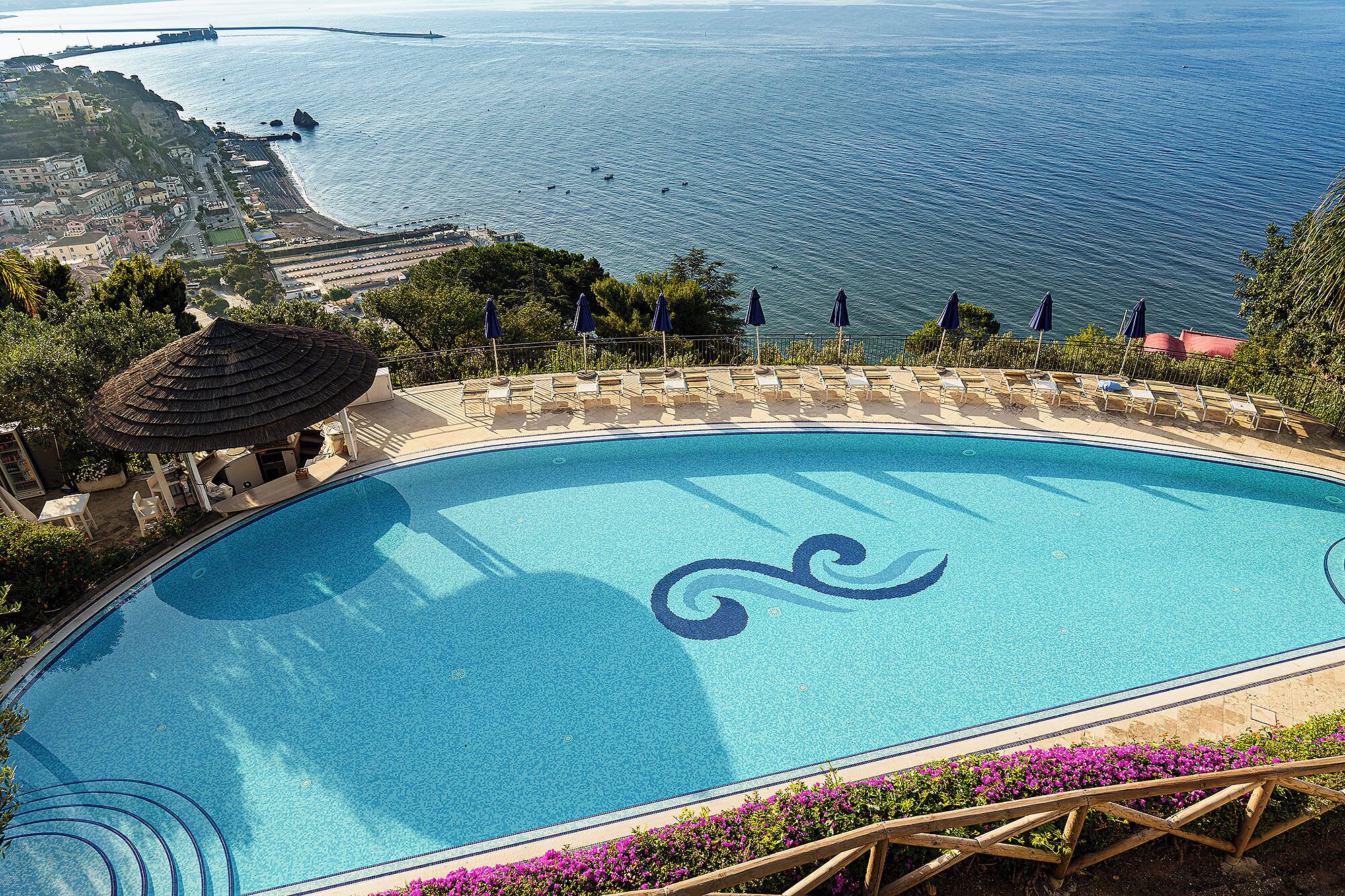Ein Schwimmbecken mit dekorativem Wellenmuster, umgeben von Liegestühlen und Blick auf das Meer.