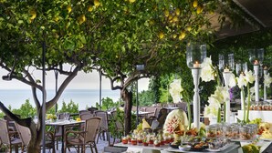 An outdoor dining area with wicker chairs and tables, a buffet with various food items, and a view of the sea.