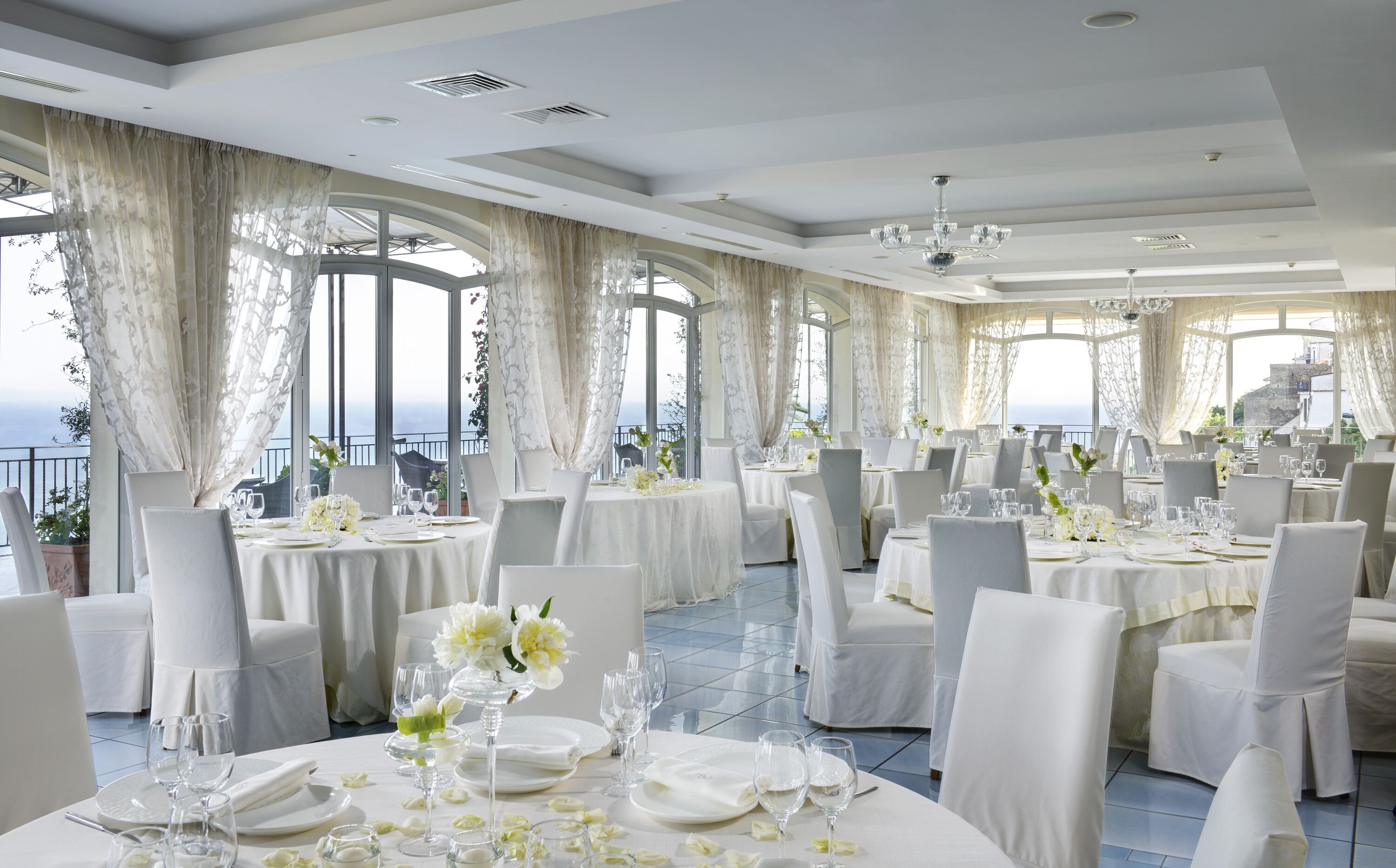 A spacious dining area with round tables, white tablecloths, and elegant chairs. Large windows offer a view of the sea.