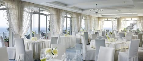 A spacious dining area with round tables, white tablecloths, and elegant chairs. Large windows offer a view of the sea.