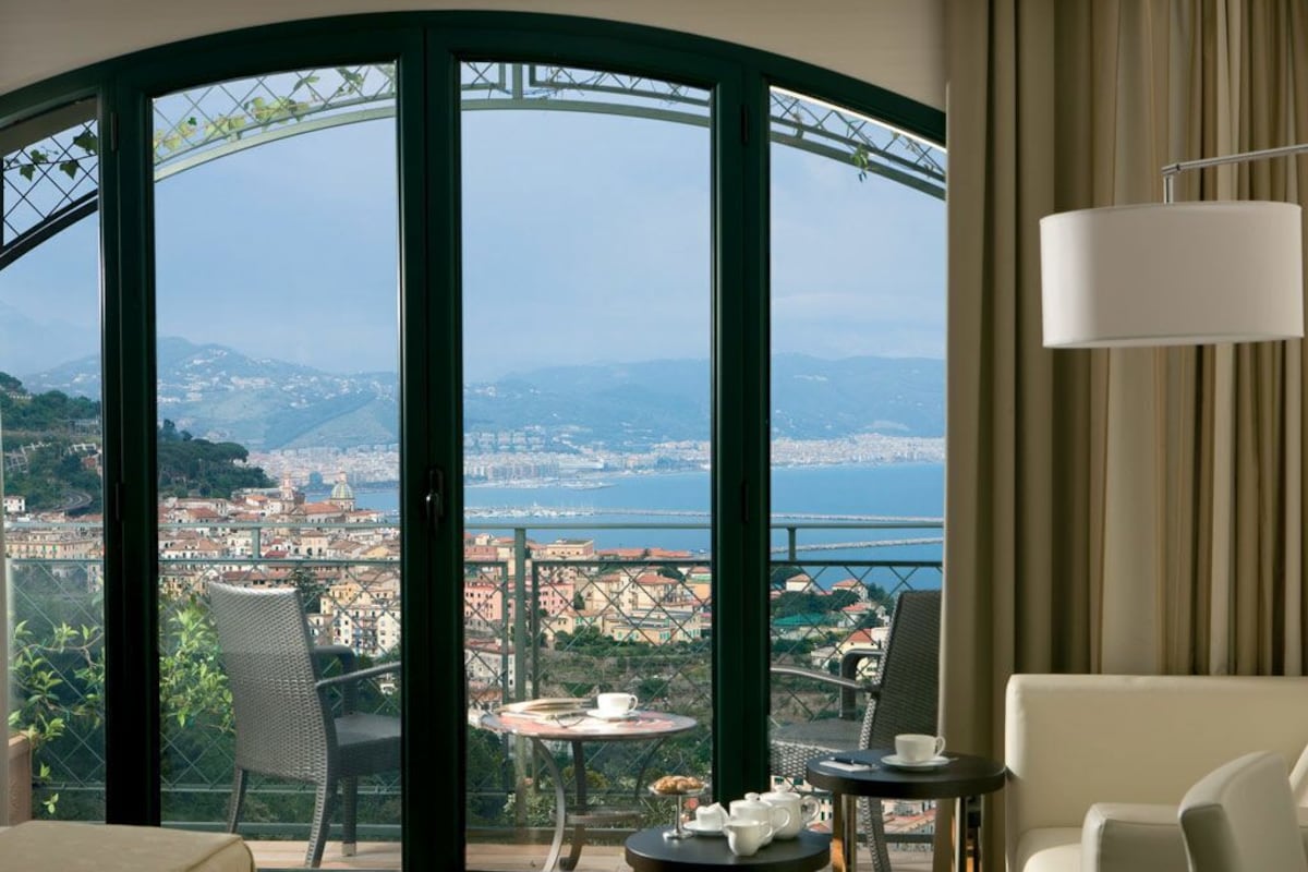 A room with a large window offering a panoramic view of a coastal city, a balcony with a table and chairs, and a sofa.