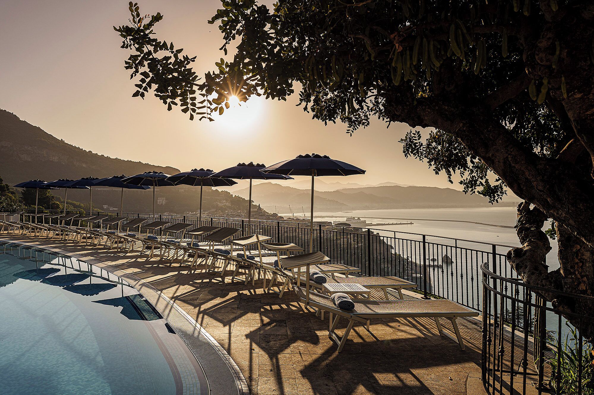 A poolside area with lounge chairs and umbrellas, overlooking a body of water and distant mountains.