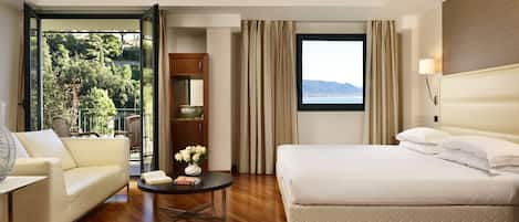 Une chambre d’hôtel avec un lit, un canapé, une petite table ornée d’un vase de fleurs, une fenêtre donnant sur des arbres et des montagnes, et un plancher en bois.