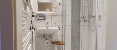 Un baño con un lavamanos blanco, un espejo, una ducha y un taburete de madera.