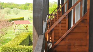 Un escalier en bois mène à un balcon d’où l’on peut admirer un champ verdoyant et un bâtiment dont le toit est rouge.
