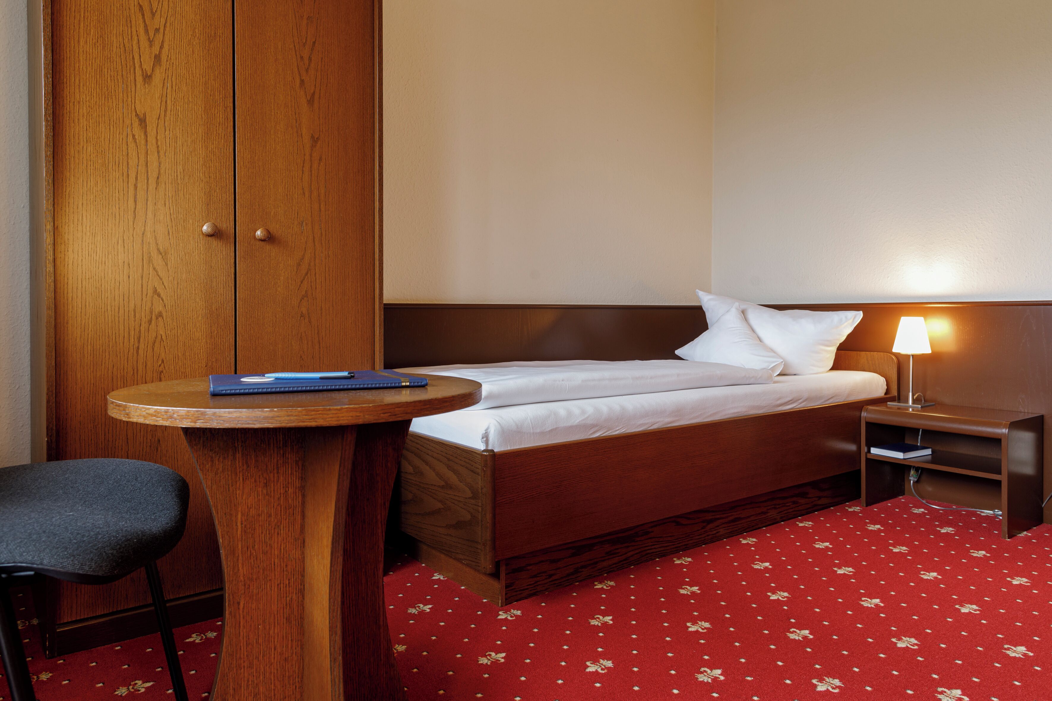 A hotel room with a wooden wardrobe, a round wooden table, a single bed with white bedding, a nightstand with a lamp, and a red floral carpet.