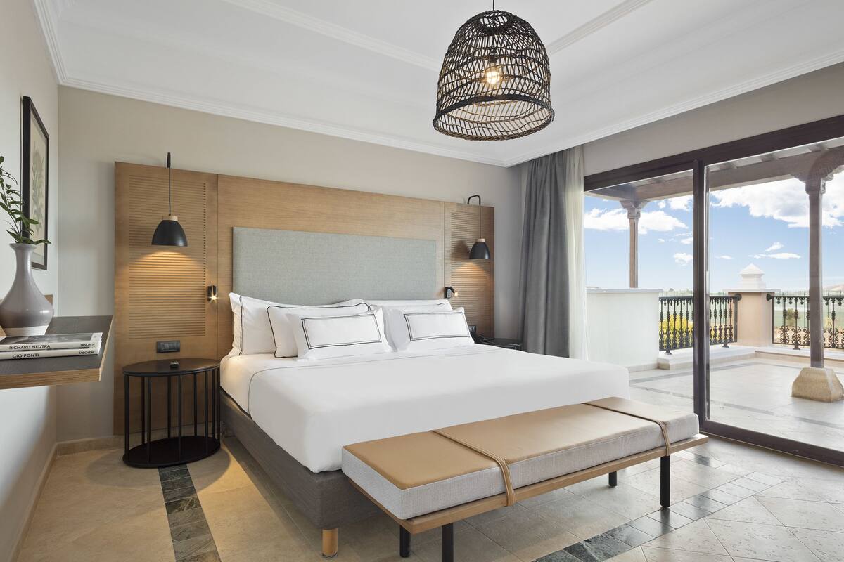 A modern hotel room with a large bed, bedside tables, a bench, and a balcony with a view.