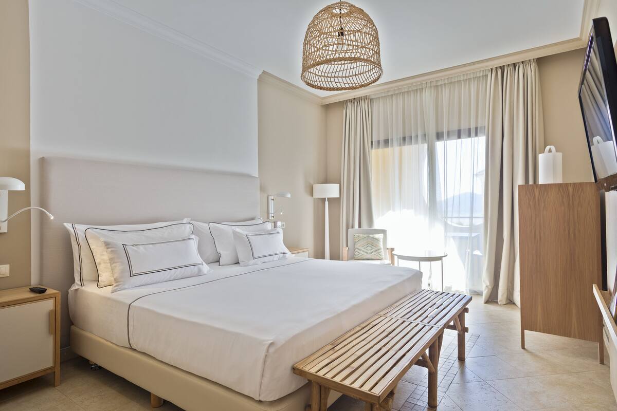 A hotel room with a large bed, bedside tables, a wooden bench, a TV, and a balcony with curtains.