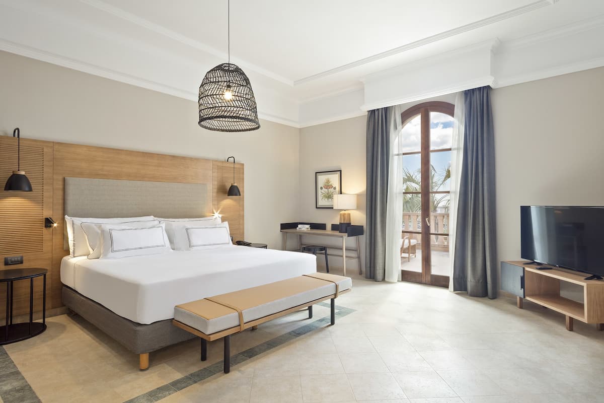 A modern hotel room with a large bed, a desk, a television, and a balcony with curtains.