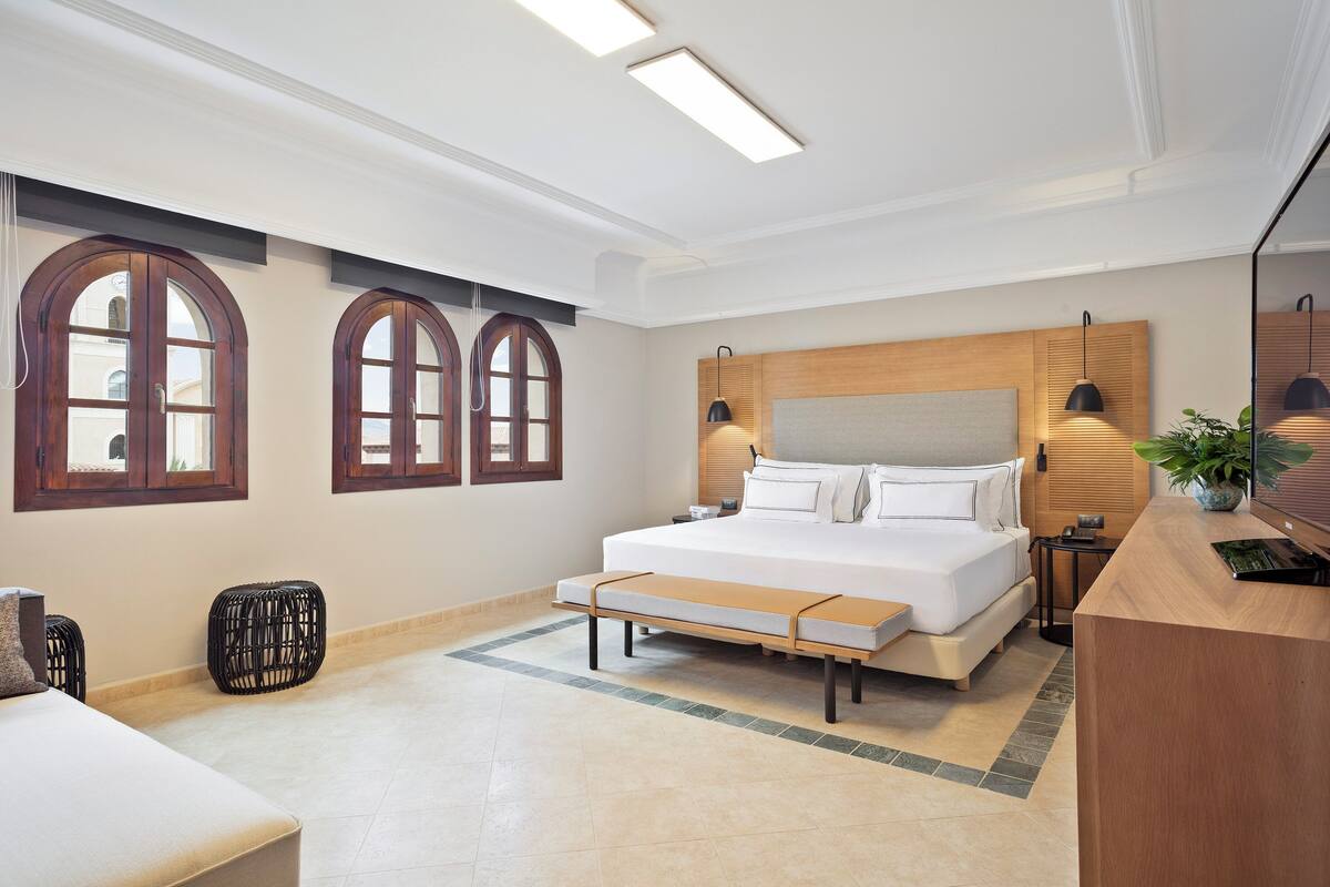 A modern bedroom with a large bed, wooden headboard, two windows with arched tops, a bedside table, and a bench.