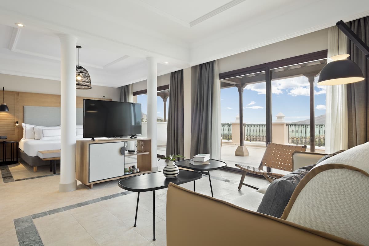 A modern hotel room with a bed, a TV, a dining area, and a balcony with a view.