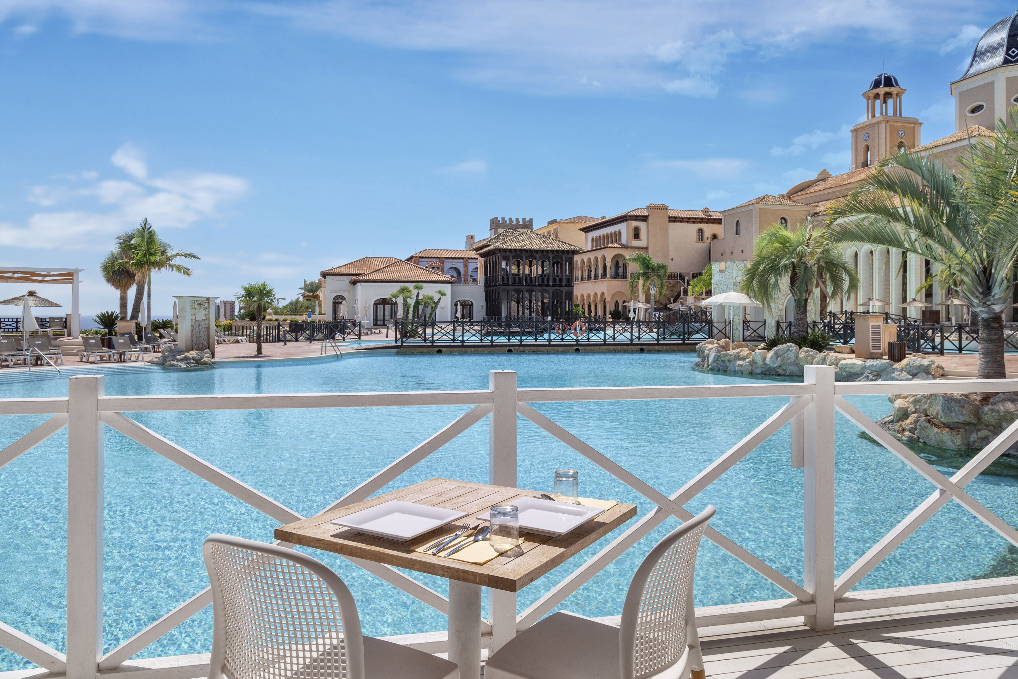 Zona de piscina con una mesa dispuesta para comer, vista despejada de la piscina y un edificio con una torre de campanas al fondo.