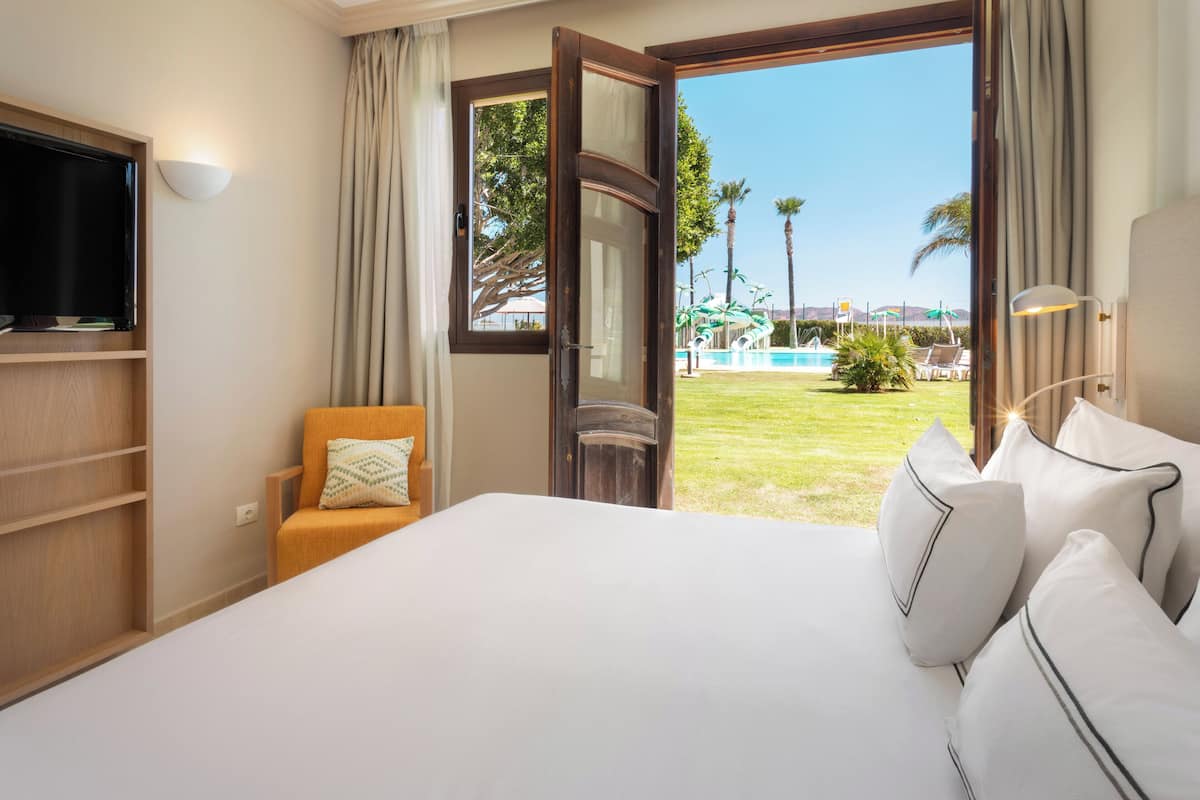 A hotel room with a bed, a TV, a chair, and a view of a swimming pool and palm trees.