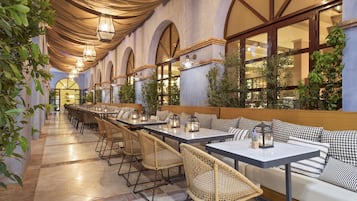 A restaurant with a long dining area, wicker chairs, and a white sofa with checkered cushions.