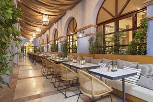 A restaurant with a long dining area, wicker chairs, and a white sofa with checkered cushions.