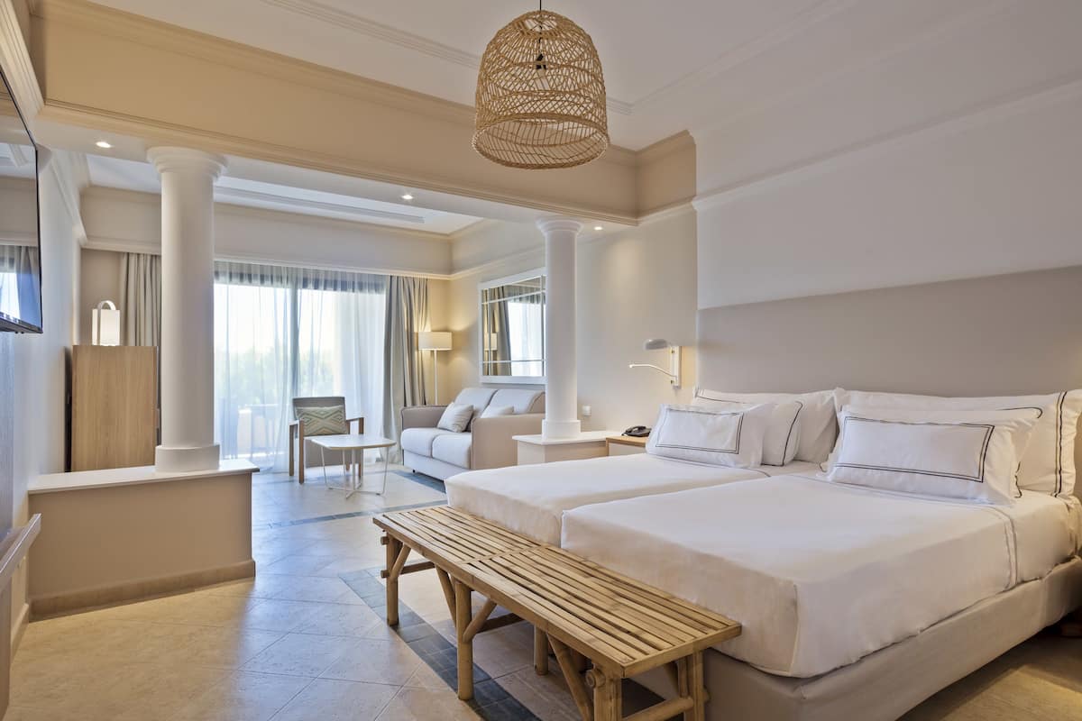 A hotel room with two beds, a wooden bench, a hanging light fixture, and a view of the outdoor area.