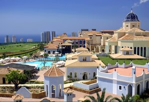 A resort with a swimming pool, multiple buildings with Mediterranean architecture, and a view of the ocean.