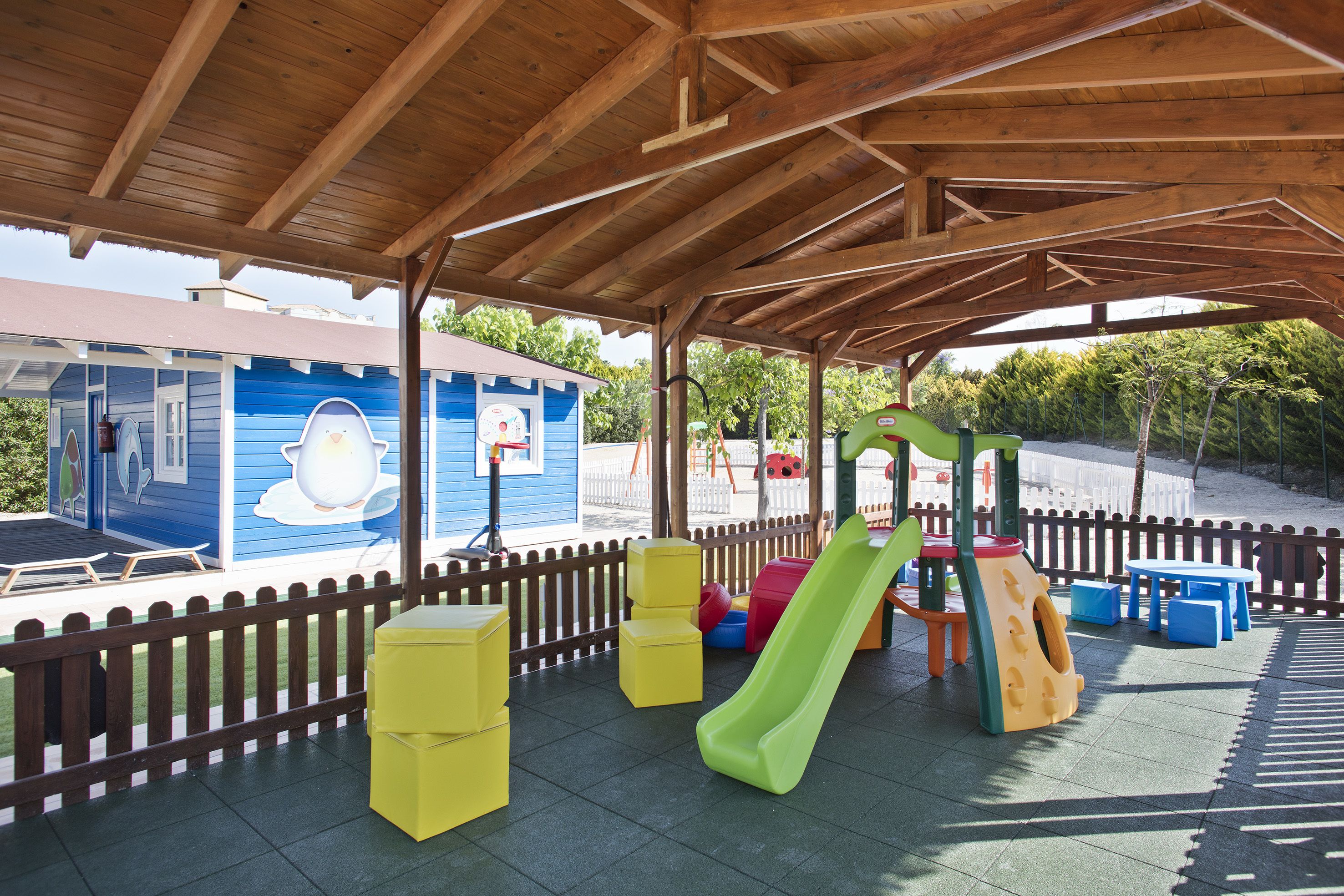 Un'area giochi coperta con uno scivolo, panchine e un edificio con pareti blu.