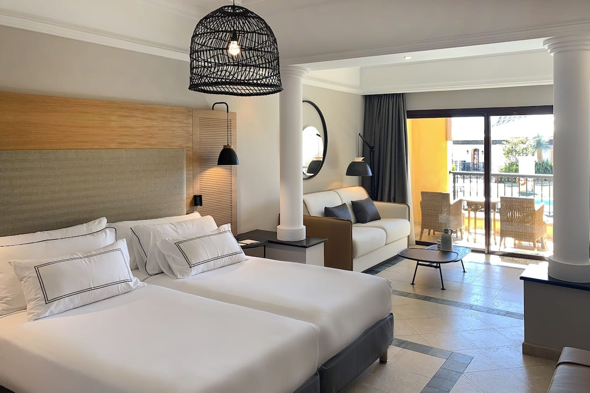 A hotel room with two large beds, a sofa, a small table, and a balcony with wicker furniture.