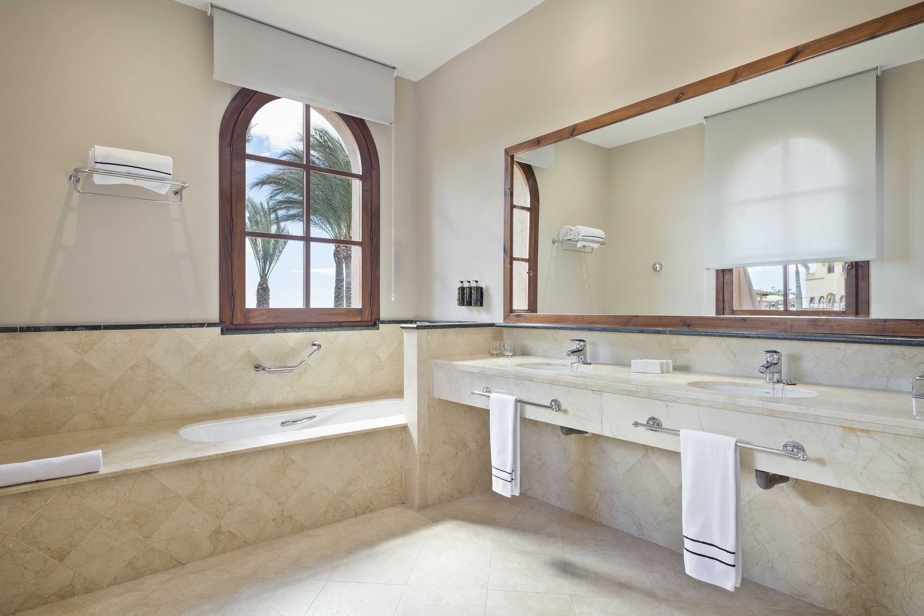 Un baño con bañera, dos lavamanos, un espejo grande y una ventana con vista a palmeras.
