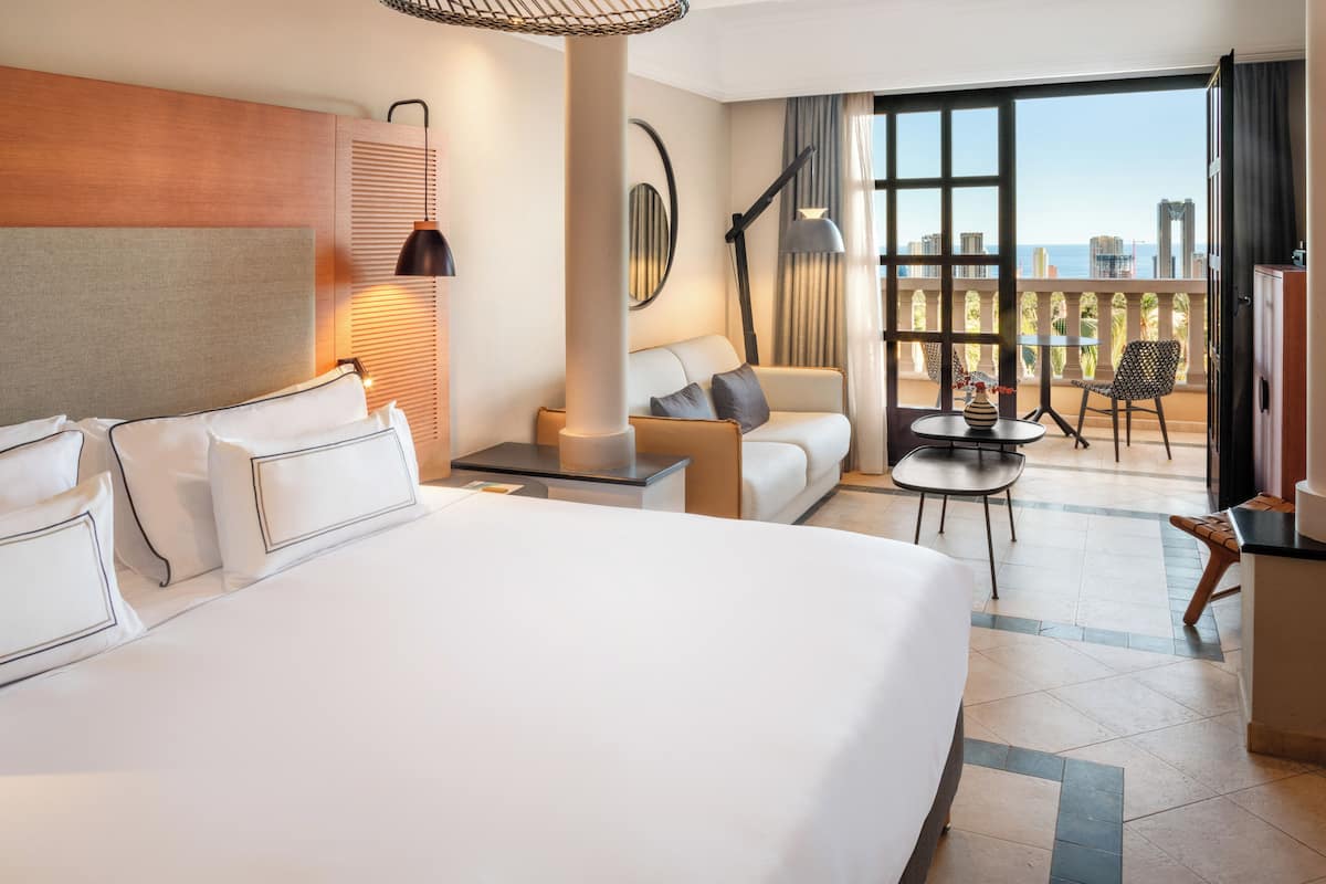 A hotel room with a large bed, a sofa, a balcony with a table and chairs, and a city view.