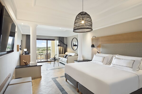 A modern hotel room with a large bed, a sitting area with a sofa and armchairs, a dining table, and a balcony with a view.