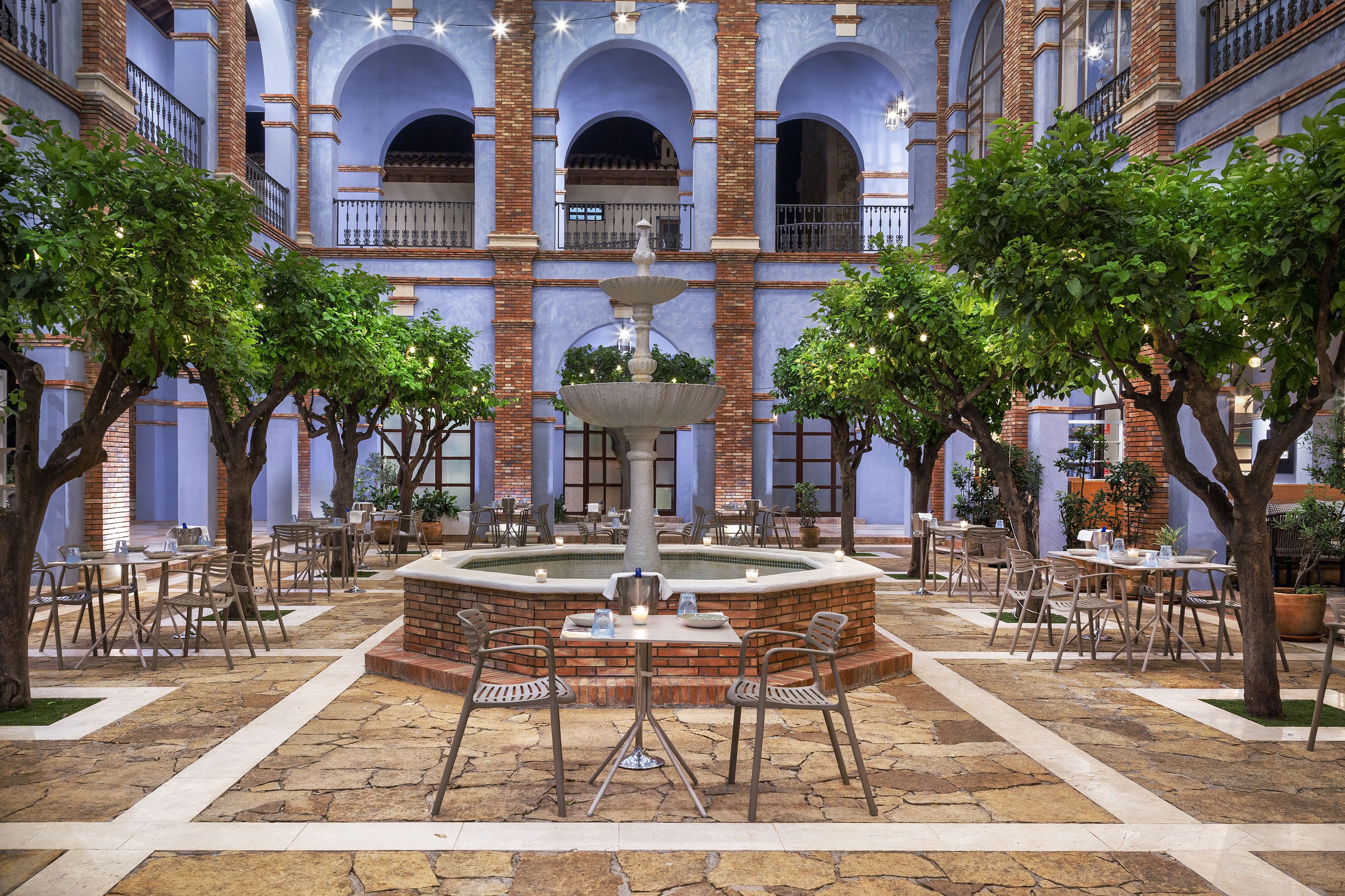 Un pati interior amb una font central, envoltat de taules i cadires, i un edifici de maó amb arcs al fons.
