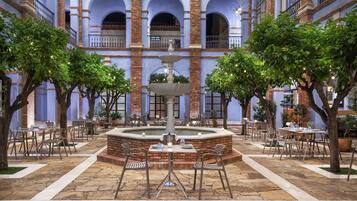 Un pati interior amb una font central, envoltat de taules i cadires, i un edifici de maó amb arcs al fons.